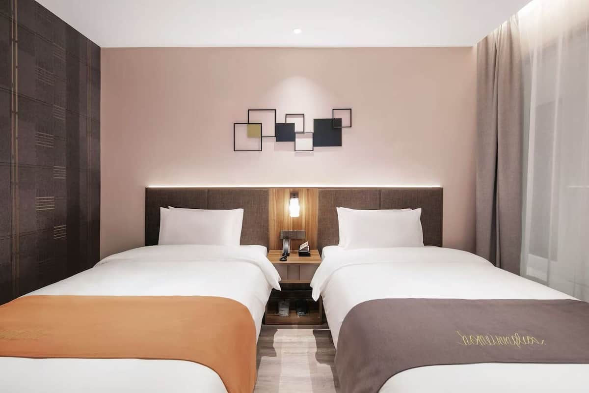 A hotel room with two beds, a headboard with built-in lighting, a wall-mounted artwork, and a large window with curtains.