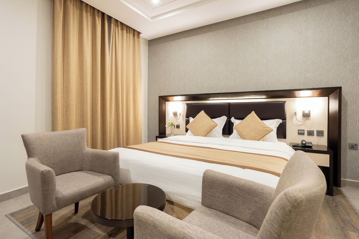A hotel room with a large bed, two armchairs, a small table, and a nightstand with a lamp.