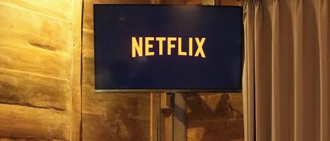 Interior de una cabaña de madera con una televisión en la pared mostrando el logotipo de Netflix, un escritorio de madera y un taburete pequeño.