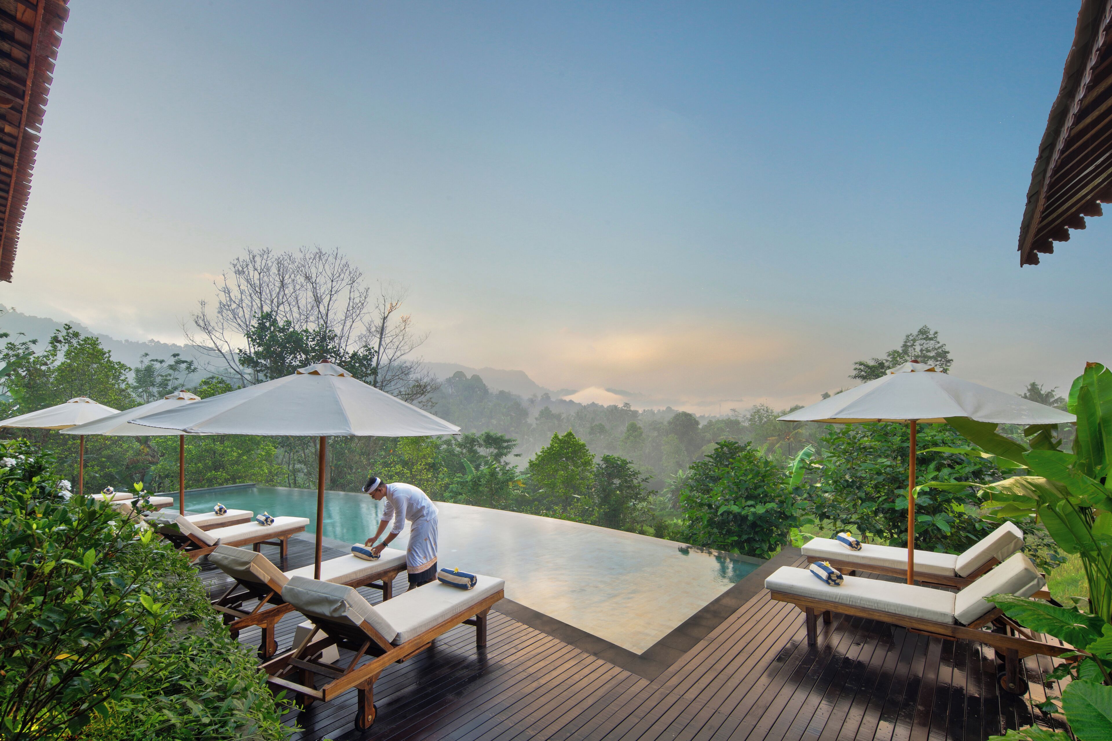 An infinity pool with lounge chairs and umbrellas, surrounded by lush greenery and mountains in the background.