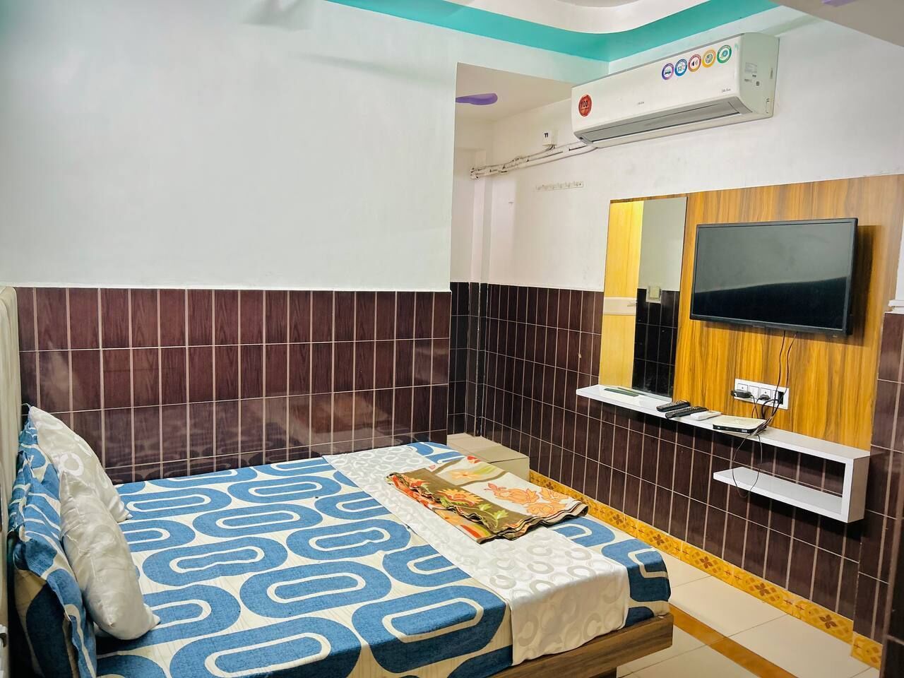 A hotel room with a bed, a TV mounted on the wall, a small desk with a chair, and an air conditioning unit above the bed.