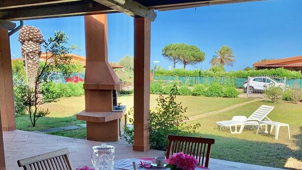 Un patio al aire libre con una mesa dispuesta para tomar té, rodeado de sillas, una parrilla y vistas a un jardín y árboles.