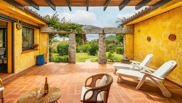 Un patio cubierto con pilares de piedra, vistas a la vegetación y mobiliario exterior.