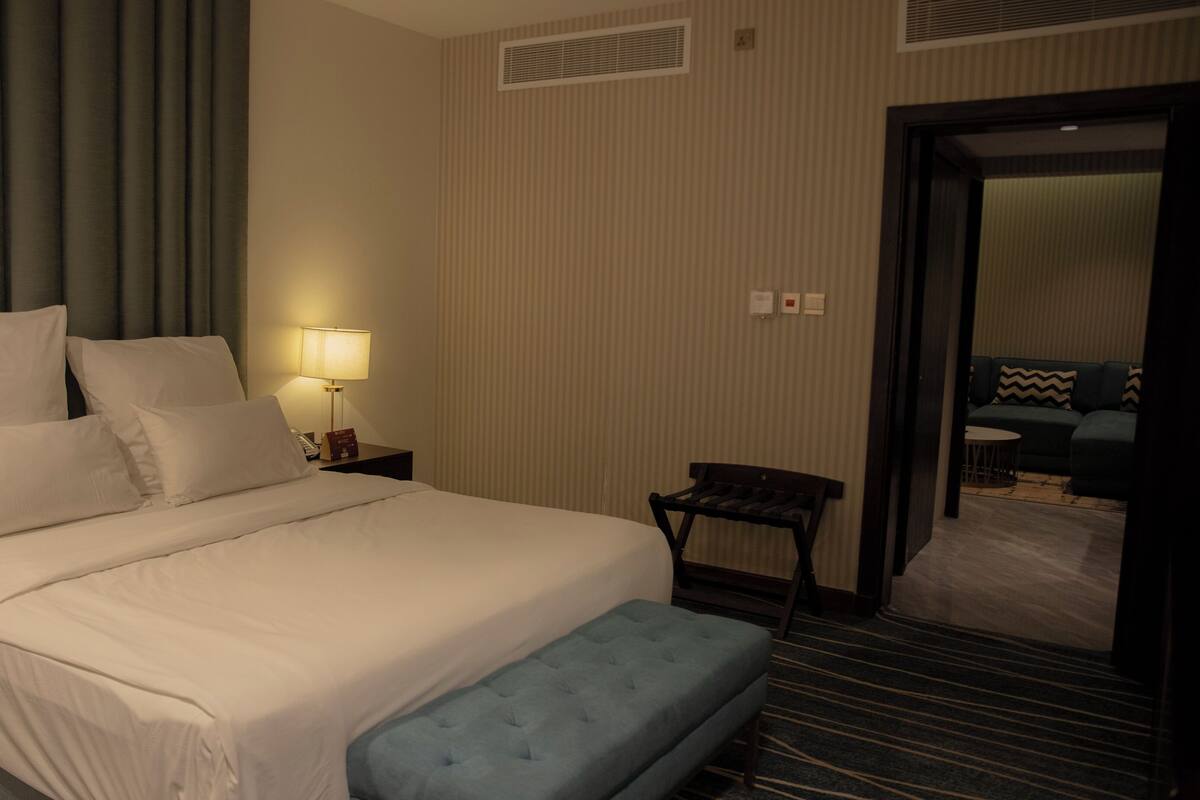 A hotel room with a large bed, a bedside table with a lamp, a small table, and a bench.