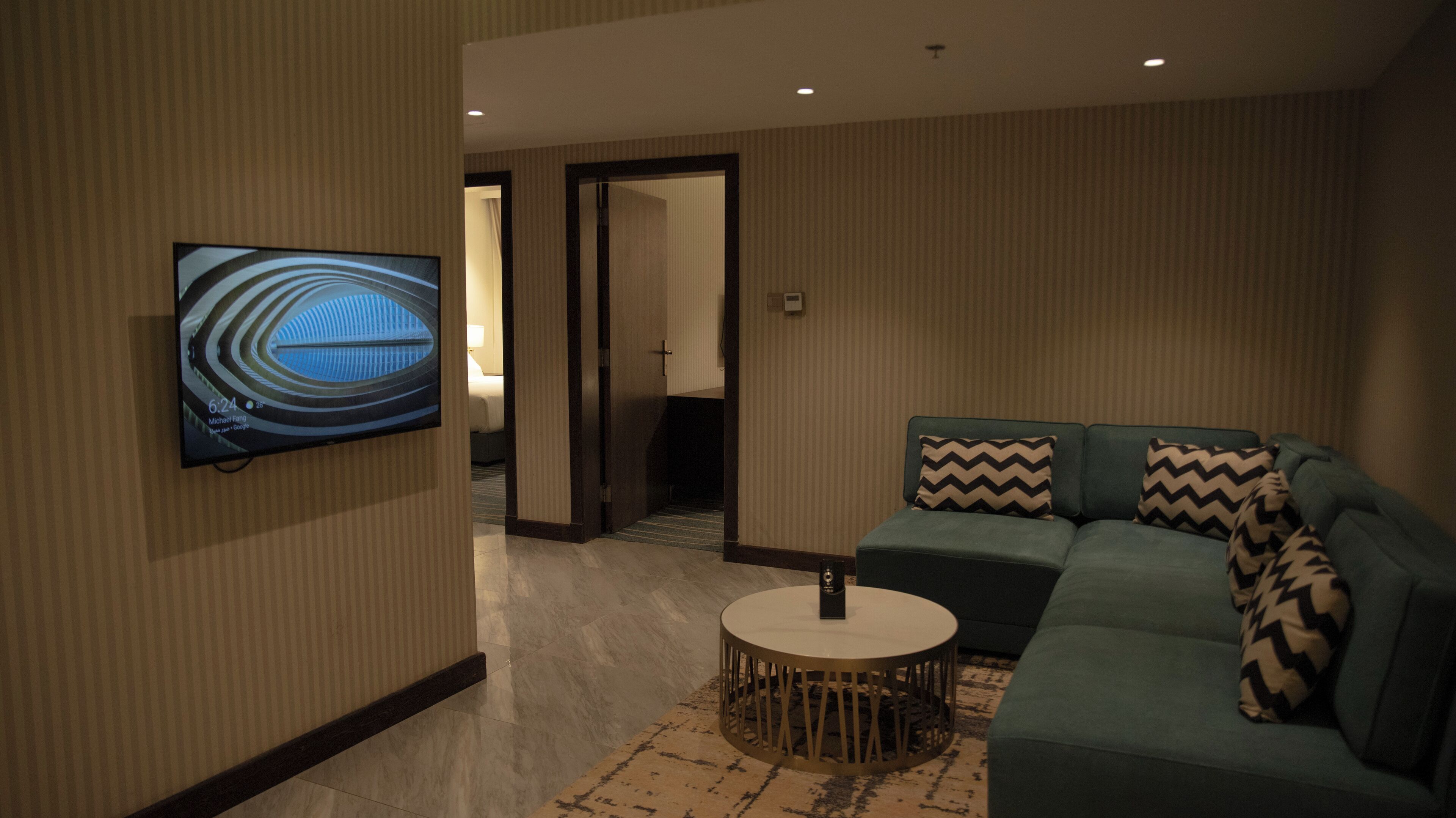 A hotel room with a green sofa, a round coffee table, and a flat-screen TV mounted on the wall.