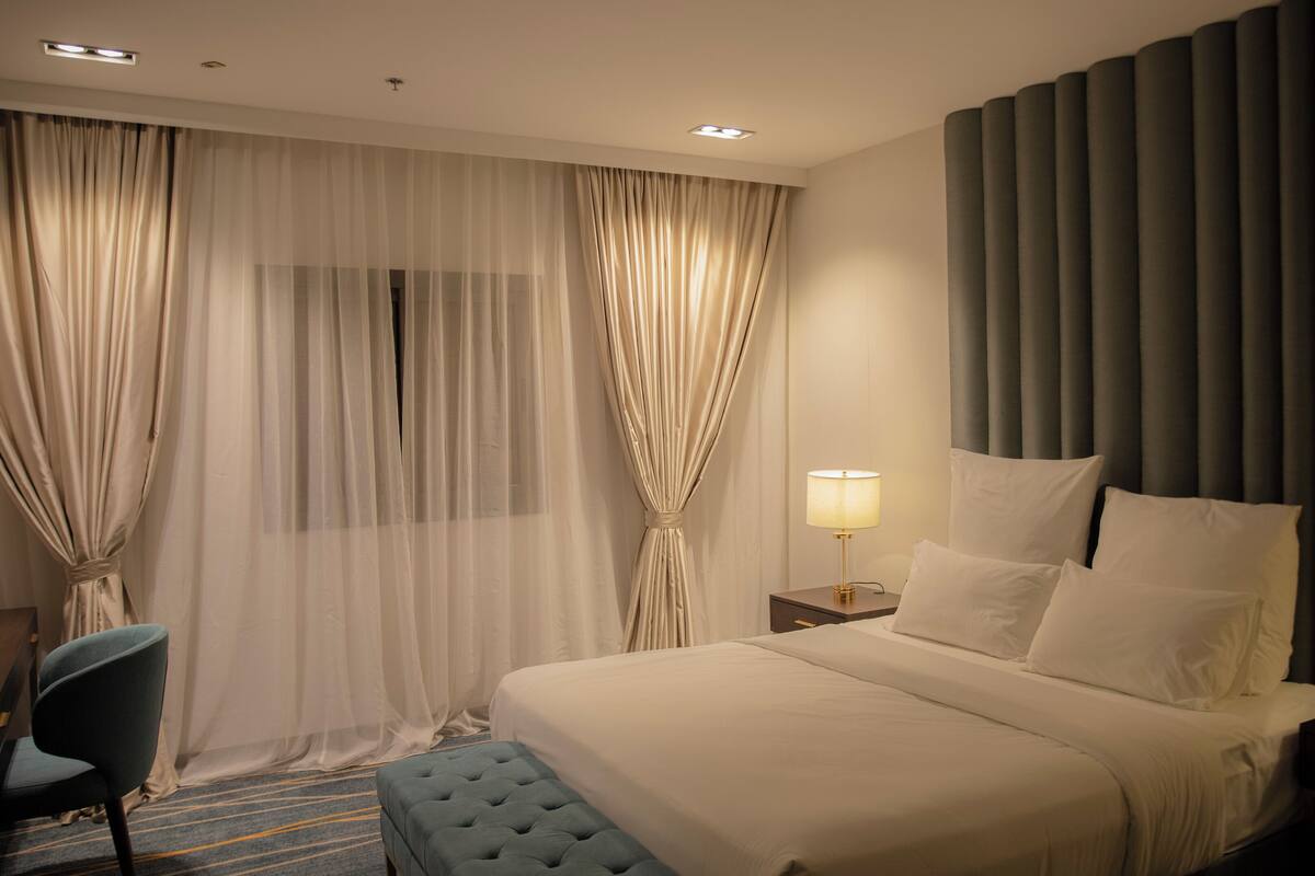 A hotel room with a large bed, a nightstand with a lamp, a chair, and sheer curtains.