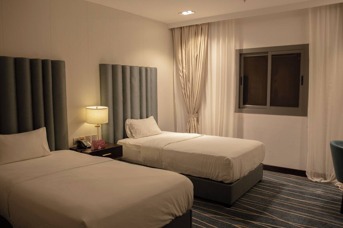 A hotel room with two beds, a nightstand with a lamp, a window with curtains, and a chair.