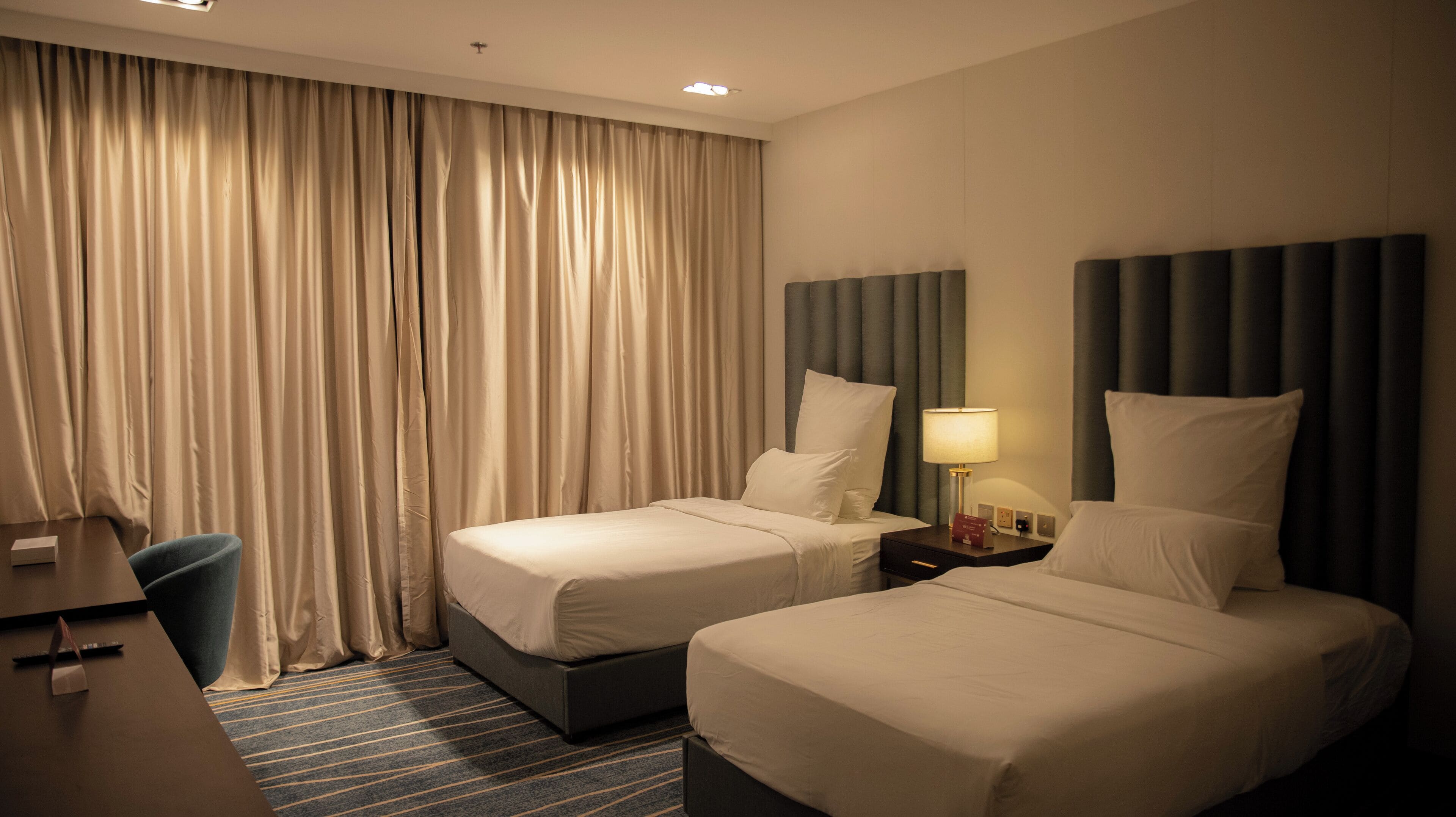 A hotel room with two beds, a desk, a chair, and a large window with curtains.