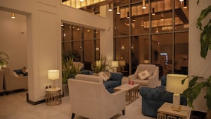 Un vestíbulo moderno con sofás, sillones y una mesa de centro. Se observan amplias ventanas y una escalera al fondo.