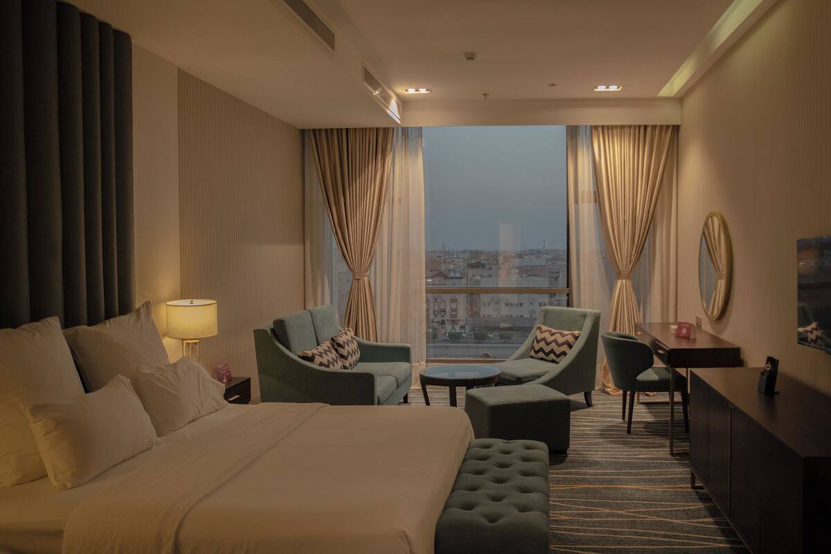 A hotel room with a large bed, two armchairs, a small table, a TV, and a view of the city through large windows.