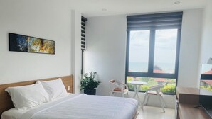Un dormitorio moderno con una cama grande, un escritorio, una silla, una planta en maceta y vistas al mar a través de un ventanal amplio.