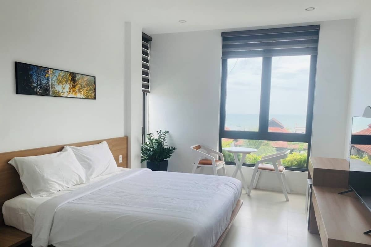 Ein modernes Schlafzimmer mit einem großen Bett, einem Schreibtisch, einem Stuhl, einer Topfpflanze und Meerblick durch ein großes Fenster.