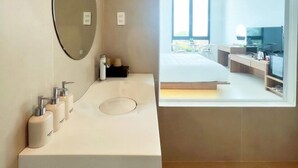 Un baño moderno con un espejo grande, un lavamanos blanco y una ventana con vistas a un dormitorio.