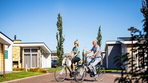 Two people riding bicycles on a paved path in a residential area with modern houses and a clear sky.