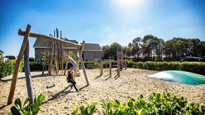 A sandy outdoor area with a swing set and a slide, surrounded by greenery and a building with a sloped roof. - Holiday Home Bemelen near Nature Reserve (Bemelen)