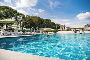 Une piscine entourée d’arbres, avec des chaises longues et des parasols, sous un ciel dégagé.