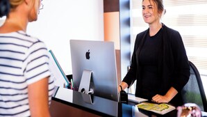 Henkilö istuu työpöydän ääressä, ja hänen edessään on kannettava tietokone ja tabletti. Huoneessa on ikkuna ja sälekaihtimet.