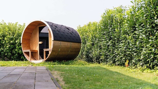 Een cilindervormige houten constructie met een open ingang, omgeven door een verzorgde haag en een verhard pad.