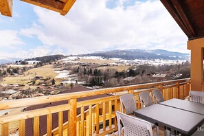 A wooden deck with a table and chairs, overlooking a snowy landscape with mountains in the distance.