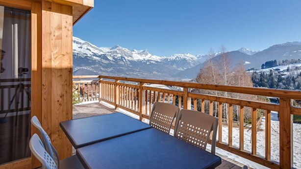 Une terrasse en bois avec une table et des chaises, offrant une vue sur des montagnes enneigées.