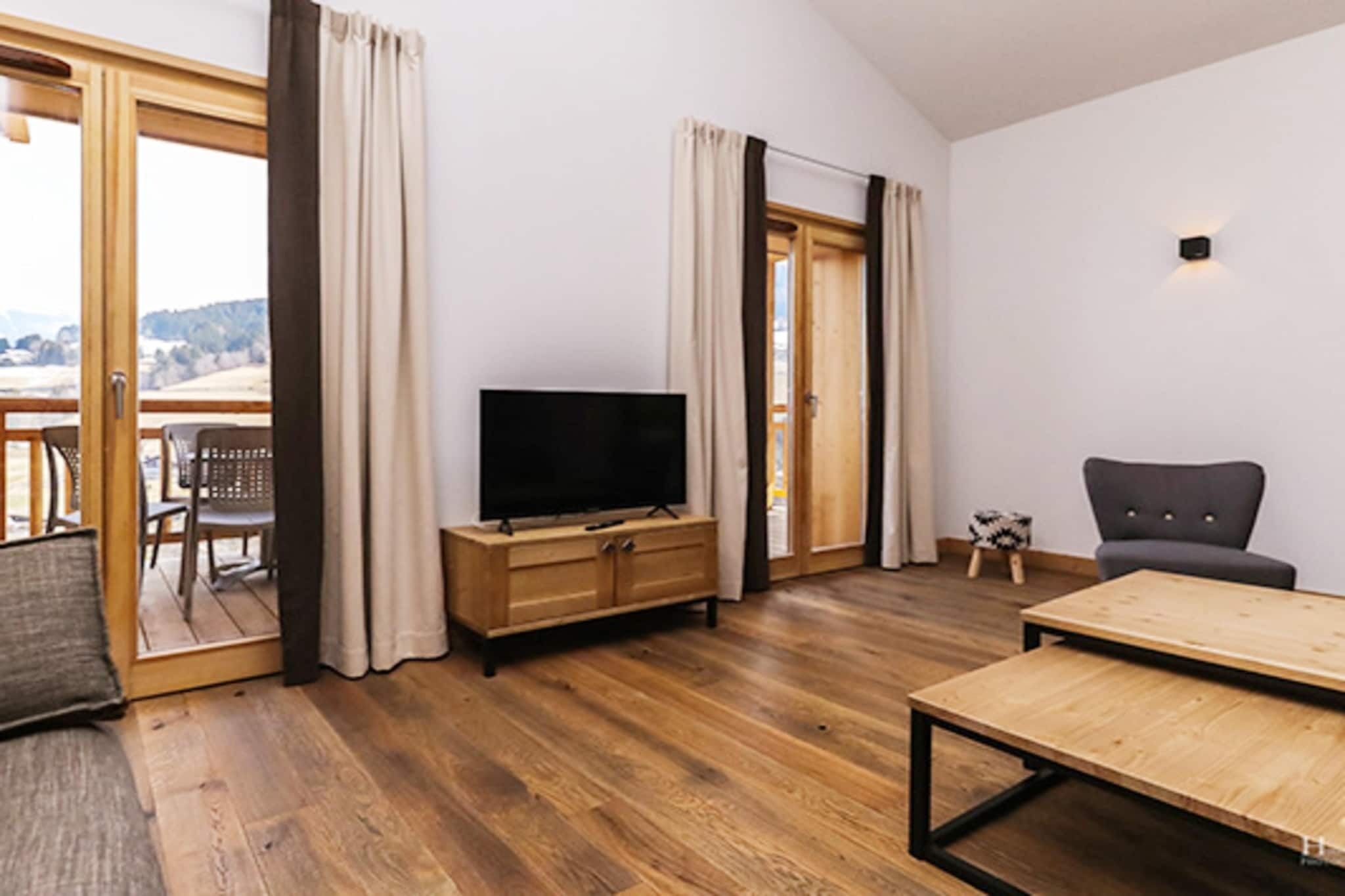 A spacious living room with a wooden floor, a flat-screen TV on a wooden cabinet, a grey sofa, and a wooden coffee table.