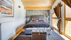 A bedroom with a large bed, a headboard, a nightstand, a lamp, a window with curtains, and a painting on the wall.