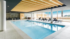 A modern indoor swimming pool with clear blue water, surrounded by lounge chairs and a stone wall.