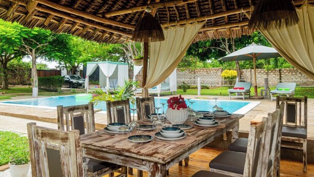 An outdoor dining area with a rustic wooden table set for four, surrounded by chairs. A pool is visible in the background, and the area is covered by a thatched roof with hanging lamps.
