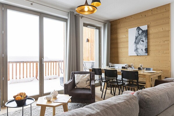 Ein modernes Wohnzimmer mit Sofa, Sessel und Esstisch. Ein großes Fenster bietet Blick auf eine Holzterrasse.