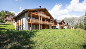 Un chalet grande de madera con varios balcones y fachada de piedra, rodeado de abundante vegetación y montañas.