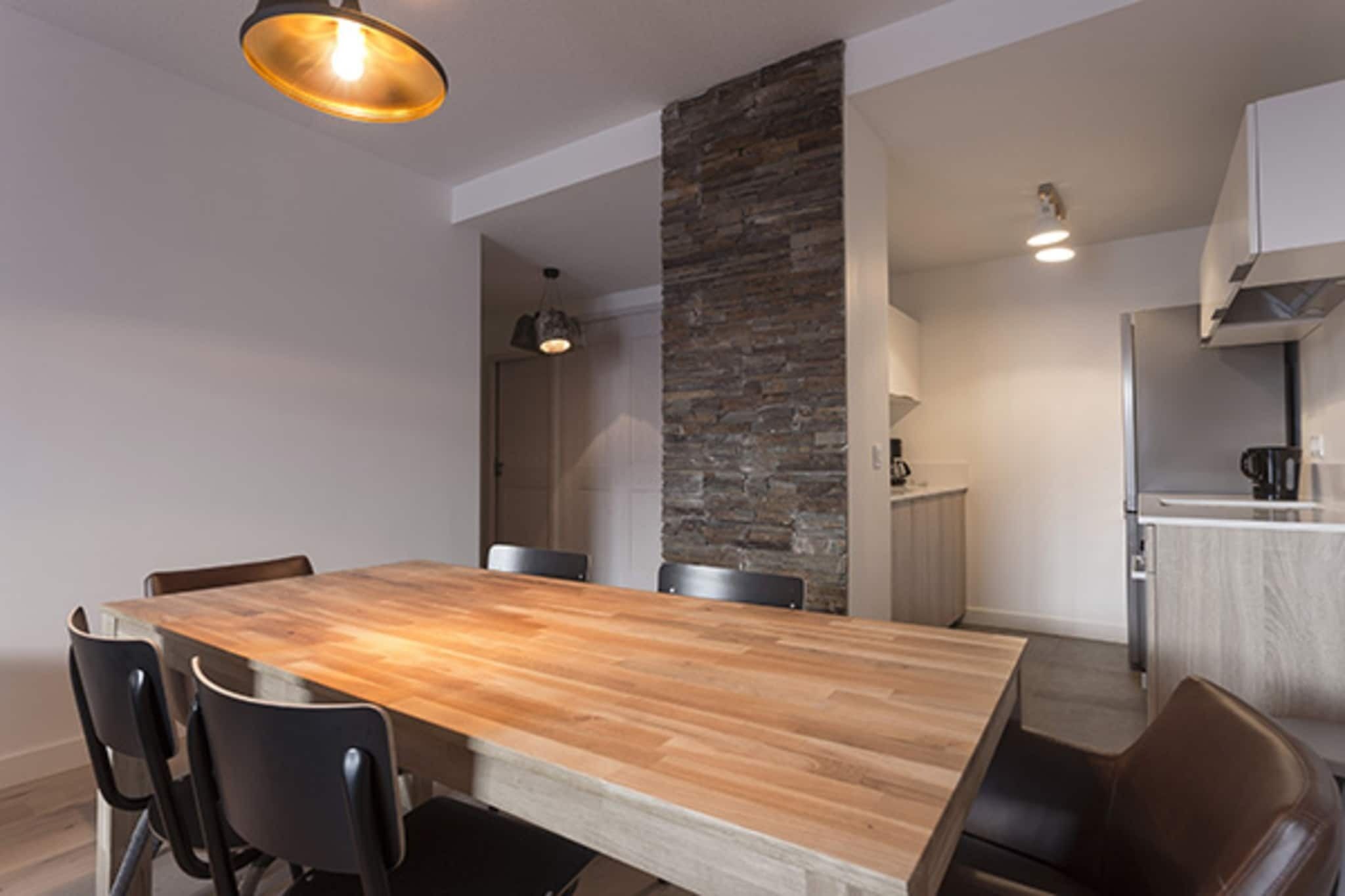 Uma área de jantar moderna com uma mesa de madeira e cadeiras pretas, uma parede de destaque em pedra e uma cozinha ao fundo.