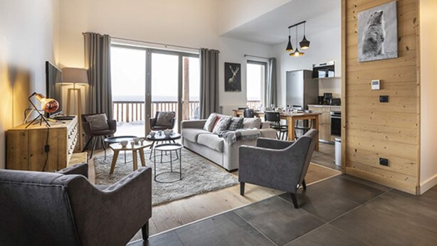 A modern living room with a sofa, chairs, a coffee table, and a dining area. There is a kitchen in the background, a balcony with curtains, and a wooden wall with a picture.