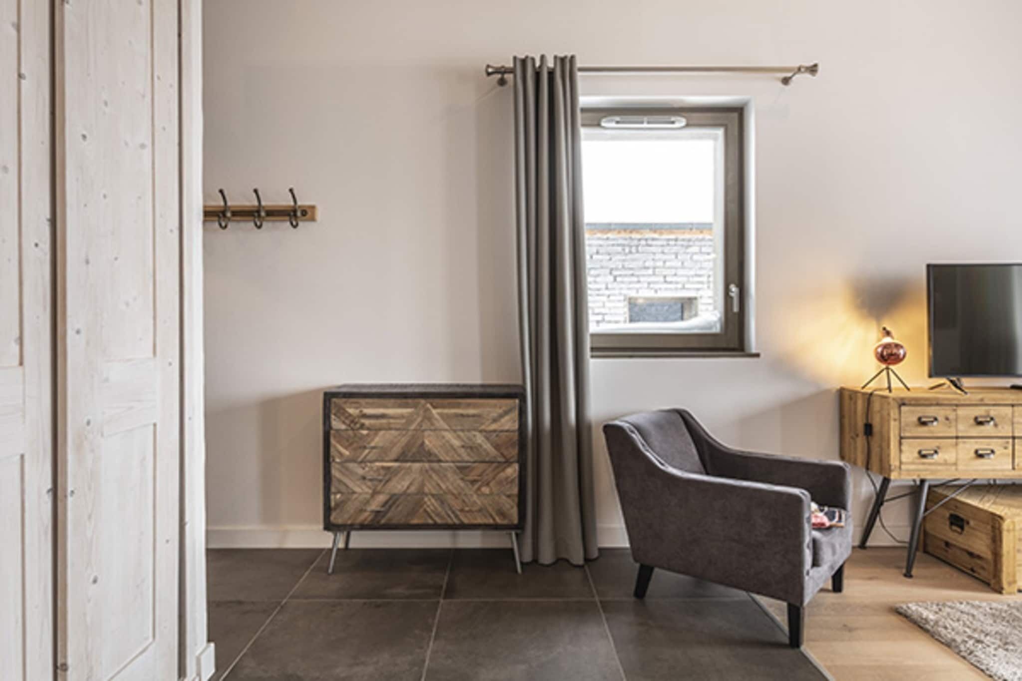 A modern living room with a grey armchair, a wooden cabinet with drawers, a radiator, a window with curtains, and a television on a wooden stand.