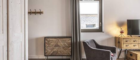A modern living room with a grey armchair, a wooden cabinet with drawers, a radiator, a window with curtains, and a television on a wooden stand.