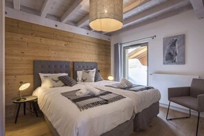 A bedroom with a wooden ceiling, a large bed with two pillows, a side table with a lamp, and a chair.
