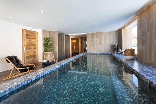 A modern indoor pool with a clear blue mosaic floor, wooden walls, and two wooden chairs.