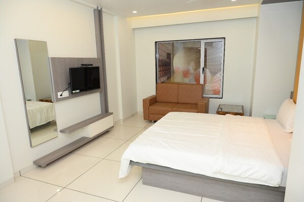 A modern hotel room with a large bed, a flat-screen TV mounted on the wall, a seating area with a brown sofa, and a large window with a view of a cityscape. - HOTEL PARQOTEL (Vapi)