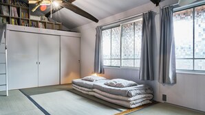 Une pièce avec un sol recouvert de tatamis, un futon servant de lit, une bibliothèque et une grande fenêtre avec des rideaux.