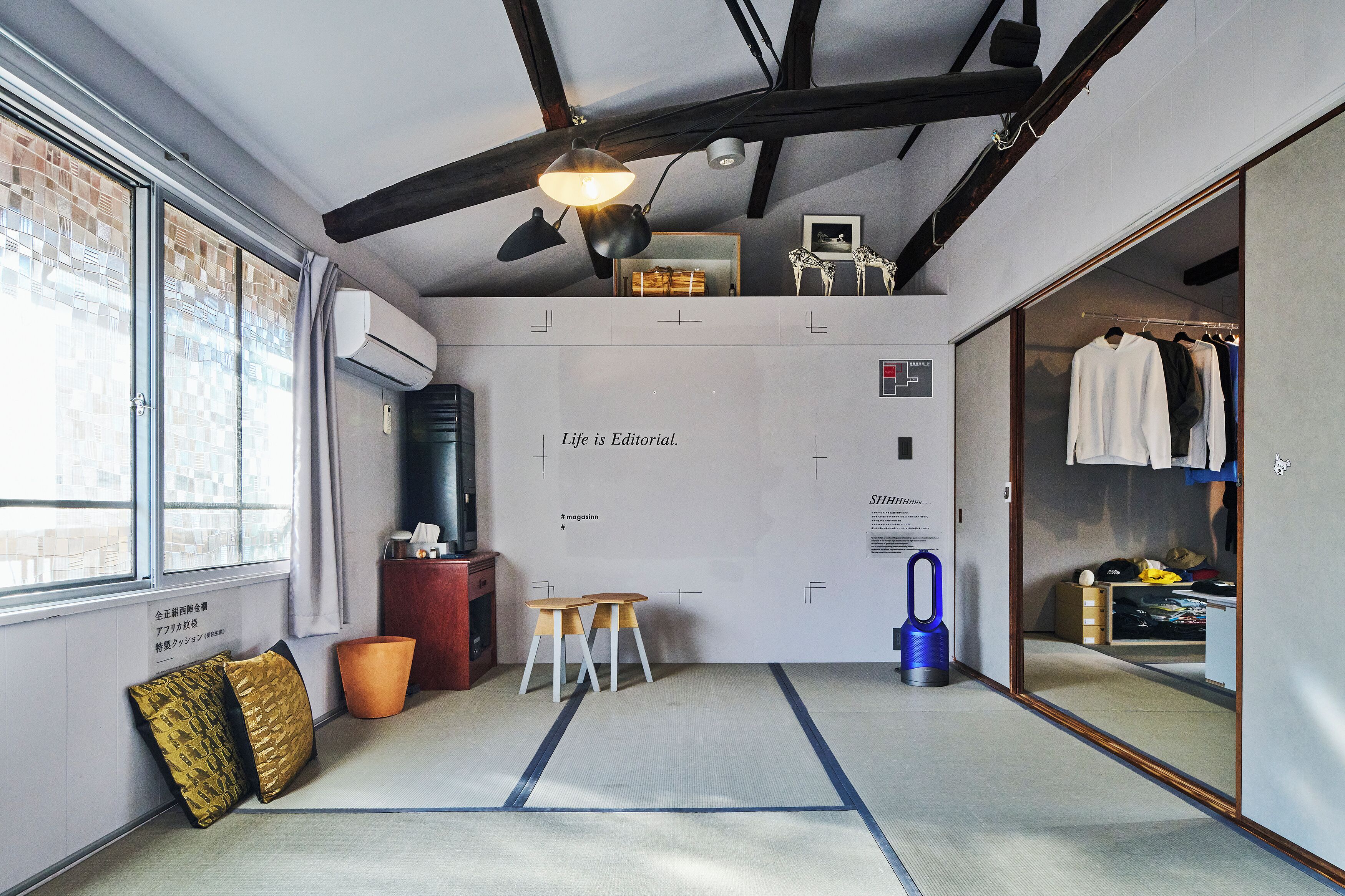 A compact room with tatami flooring, a ceiling fan, a small table with chairs, a wardrobe, and a window with a view of buildings.