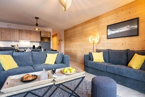 A modern living room with two grey sofas, a coffee table with a bowl of fruit and croissants, and a wooden wall with a framed picture. - Apartment in Alps with Views (AIME-LA-PLANGE)