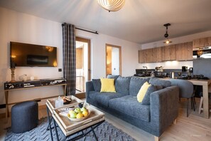 Un salon moderne avec un canapé gris, une table basse sur laquelle sont disposés des fruits, un téléviseur et un coin cuisine doté de meubles en bois.