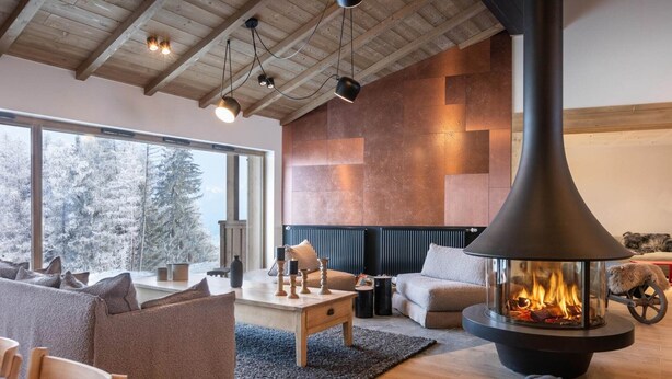 Una sala de estar acogedora con una chimenea grande, vigas de madera en el techo y vistas a árboles cubiertos de nieve.