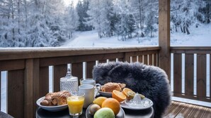Una mesa de desayuno con croissants, jugo de naranja y frutas en una terraza de madera con vistas a un bosque nevado.