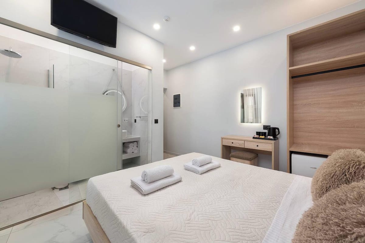 A modern hotel room with a large bed, a bedside table with a coffee maker, a mirror, and a wardrobe.
