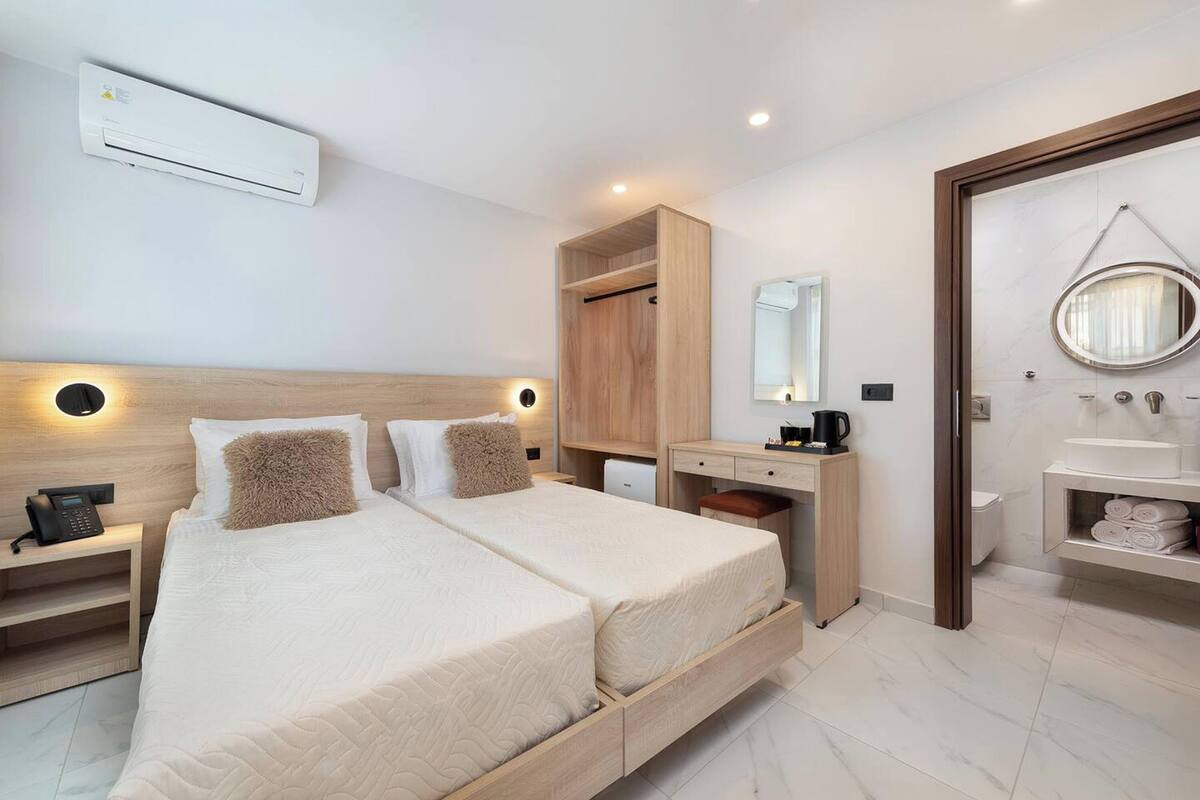 A modern hotel room with a large bed, a wooden wardrobe, a desk with a chair, a small bathroom, and a bedside table with a lamp.