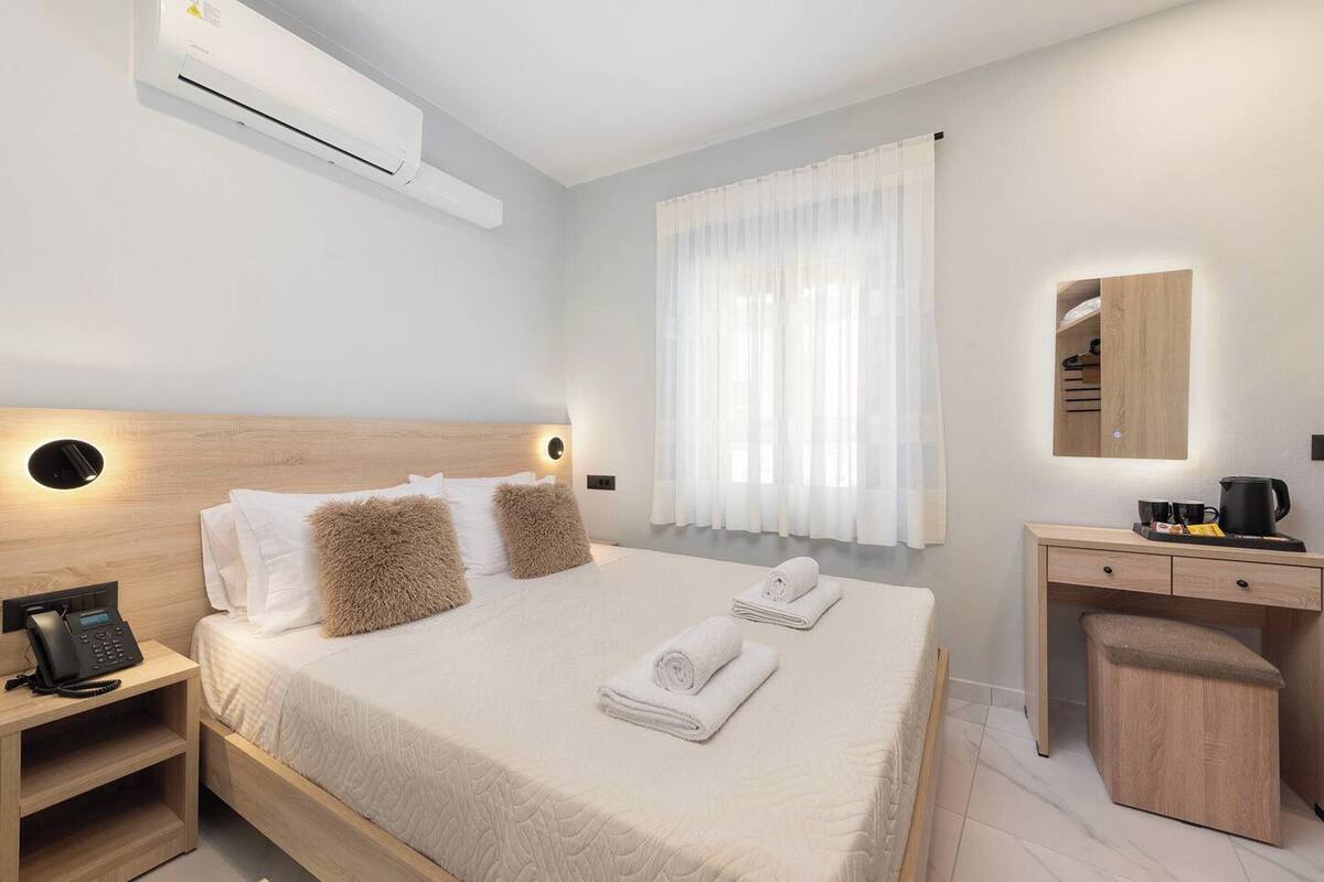 A hotel room with a bed, bedside table with a phone, a desk with a mirror, and an air conditioning unit.