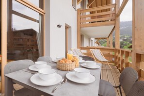 A dining table set with white plates, bowls, and a basket of bread, on a balcony with wooden railings and a view of a village.