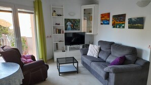 A living room with a grey sofa, a brown armchair, a glass coffee table, and a TV on a white stand. There are shelves with decorative items and a painting on the wall.