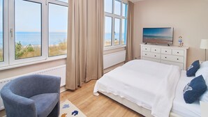 Ett sovrum med en stor säng, en stol, en tv och utsikt över havet genom stora fönster.
