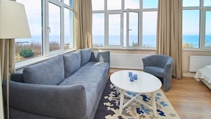 Ett modernt rum med en grå soffa, ett runt vitt bord med blå koppar och utsikt över havet genom stora fönster.